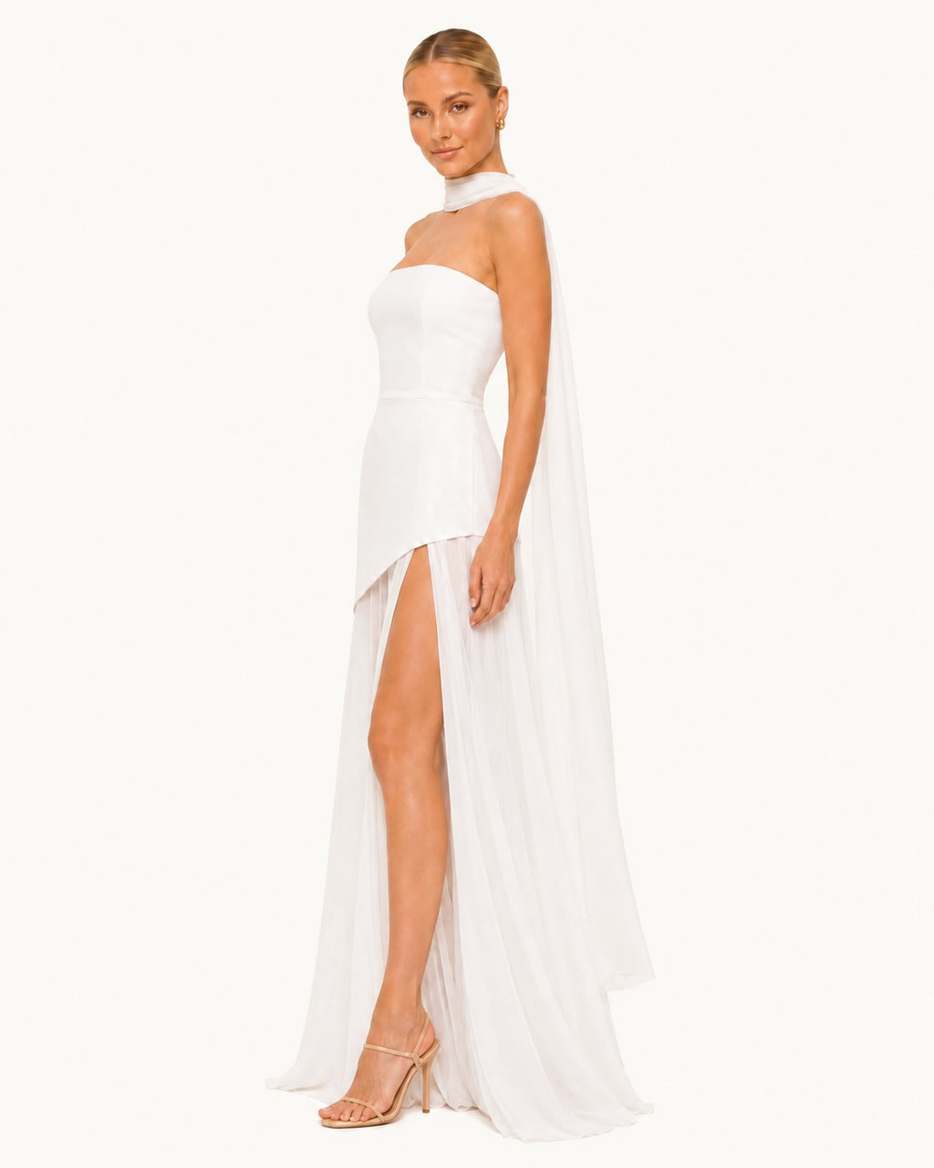 Robe de cérémonie blanche à coupe asymétrique avec fente haute et voile aérien, mettant en valeur la silhouette avec grâce et légèreté. Vue de profil.