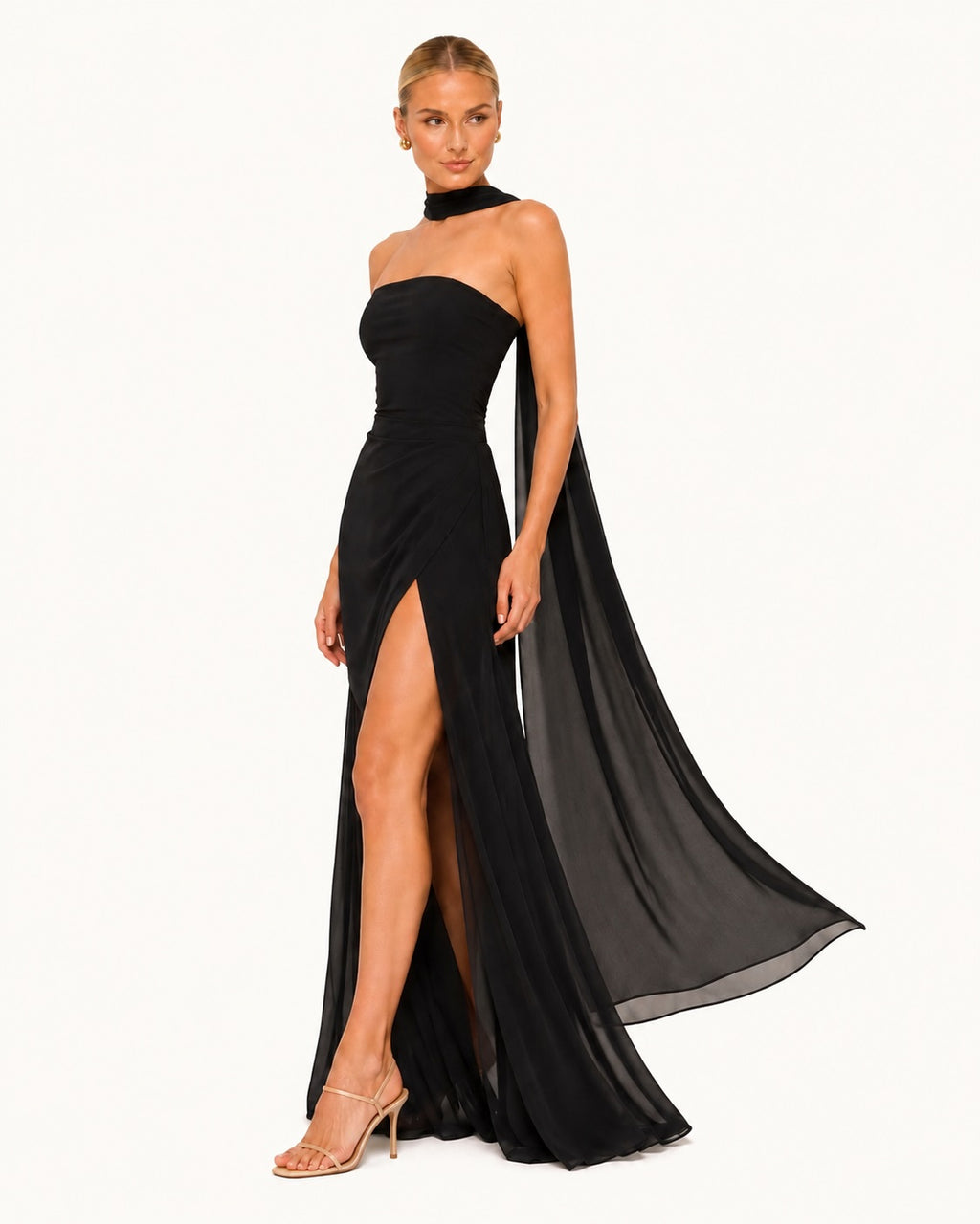 Robe de soirée noire à col foulard avec voile aérien flottant, coupe ajustée et fente sensuelle sublimant la démarche avec légèreté et raffinement. Vue trois quarts avec mouvement.