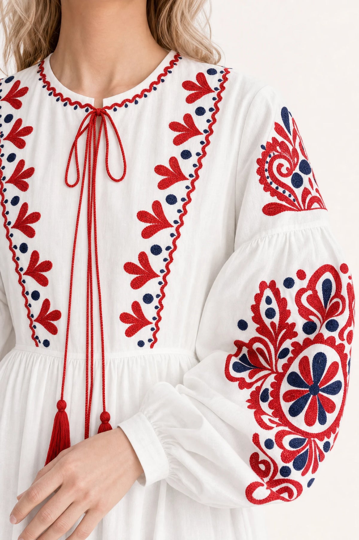 Gros plan sur la broderie bicolore rouge et bleu marine et le lien à pompons de la robe bohème brodée blanche Faustine, détail des manches bouffantes et du tissu coton