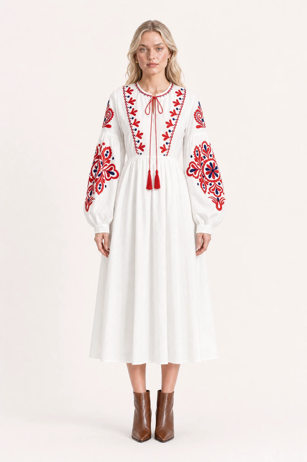 Robe bohème brodée blanche Faustine portée de face par Sophie cheveux détachés, col rond à lien pompons rouges, broderie florale bicolore rouge et bleu marine sur l'empiècement et les manches, manches longues bouffantes, bottines en cuir marron