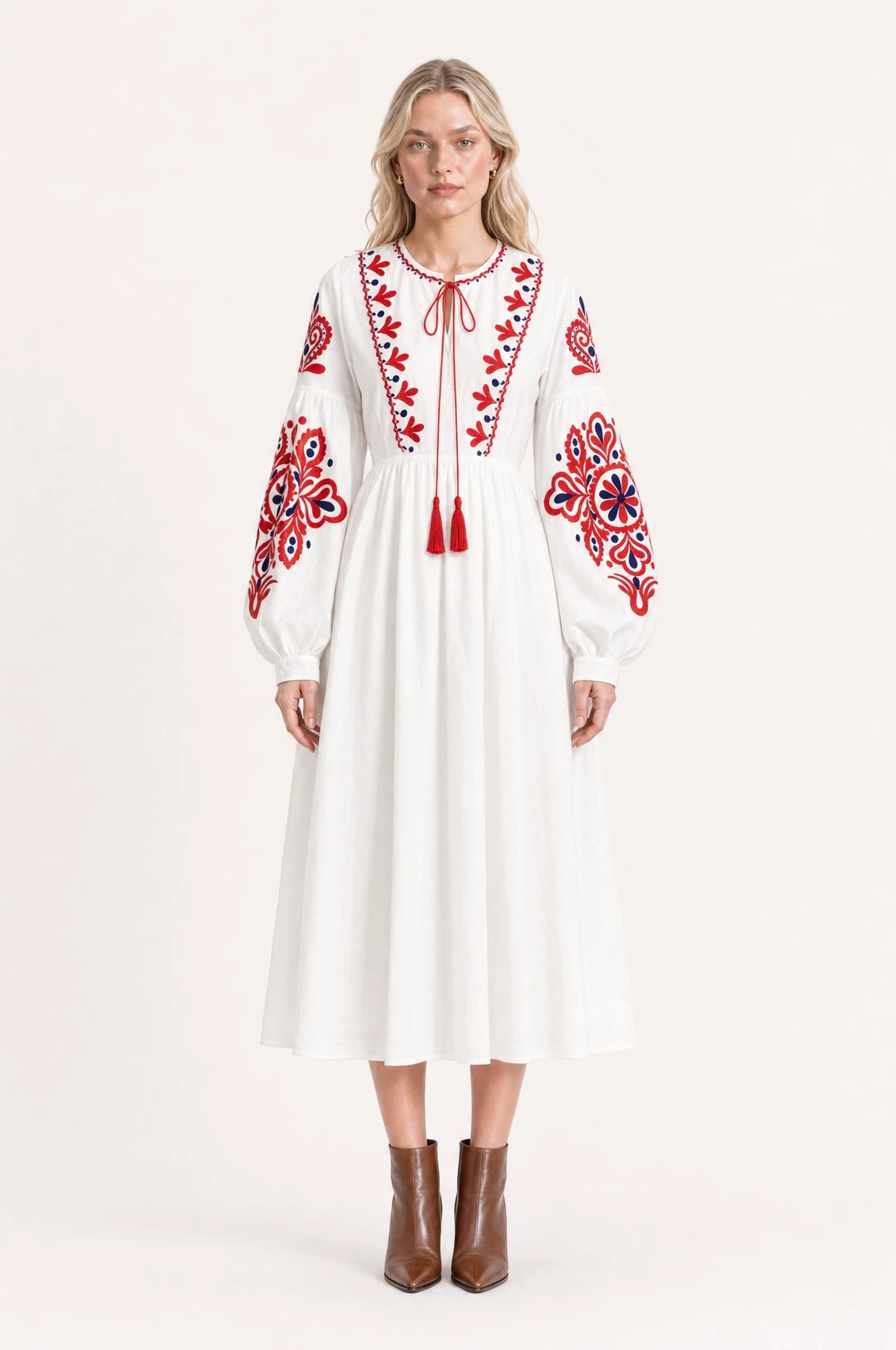 Robe bohème brodée blanche Faustine portée de face par Sophie cheveux détachés, col rond à lien pompons rouges, broderie florale bicolore rouge et bleu marine sur l'empiècement et les manches, manches longues bouffantes, bottines en cuir marron