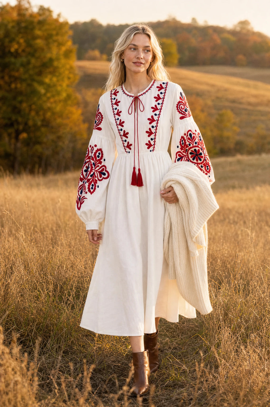 Robe bohème brodée blanche Faustine portée par Sophie dans un champ doré au coucher du soleil en automne, gilet en maille crème sur le bras, bottines marron, ambiance champêtre lumineuse