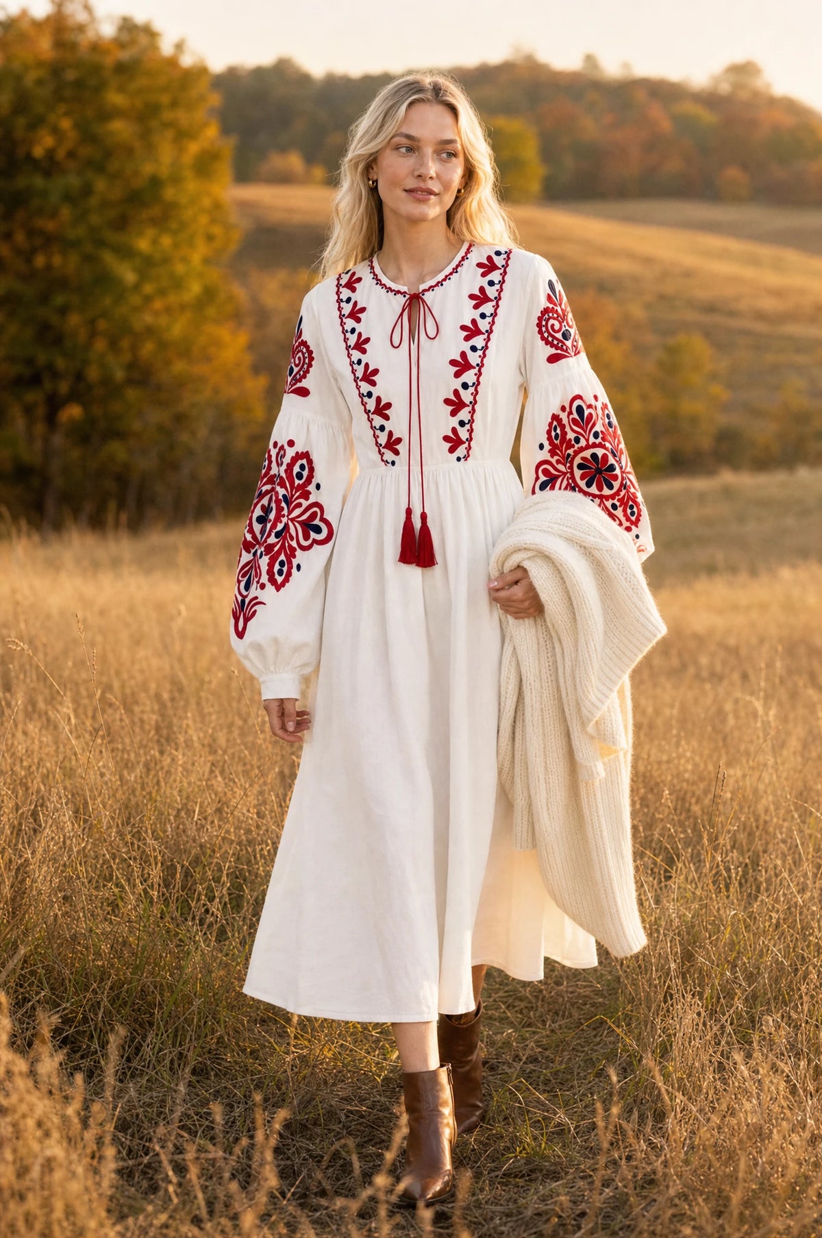 Robe bohème brodée blanche Faustine portée par Sophie dans un champ doré au coucher du soleil en automne, gilet en maille crème sur le bras, bottines marron, ambiance champêtre lumineuse Image