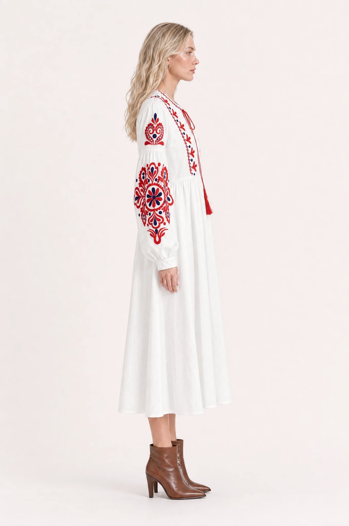 Robe bohème brodée blanche Faustine vue de profil portée par Sophie cheveux détachés, silhouette évasée taille haute, jupe plissée jusqu'à mi-mollet, bottines en cuir marron