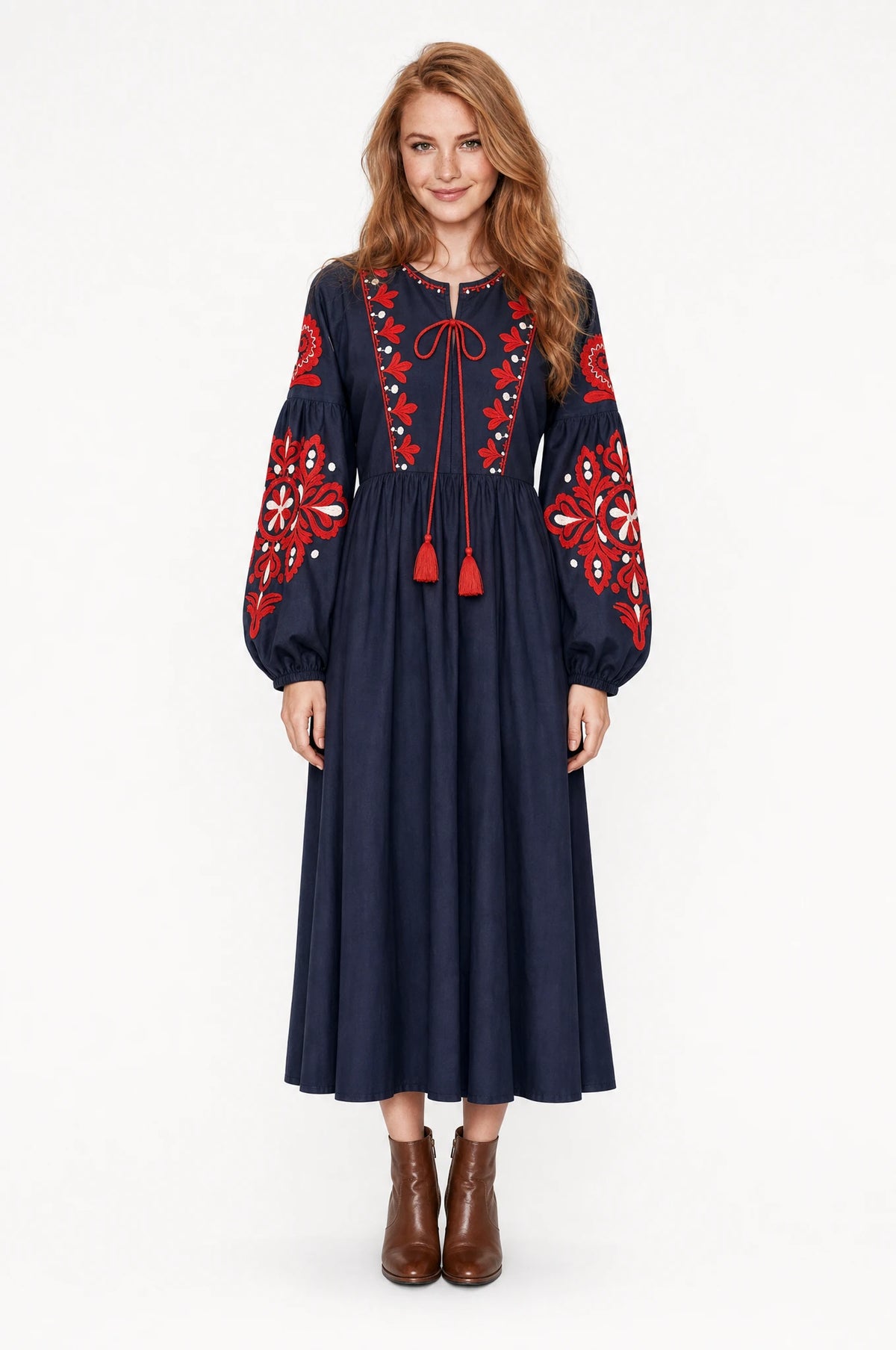 Robe bohème brodée bleu marine Constance portée de face par Julie cheveux roux ondulés détachés, col rond à lien pompons rouges, broderie florale rouge et blanche sur l'empiècement et les manches, manches longues bouffantes, bottines en cuir marron