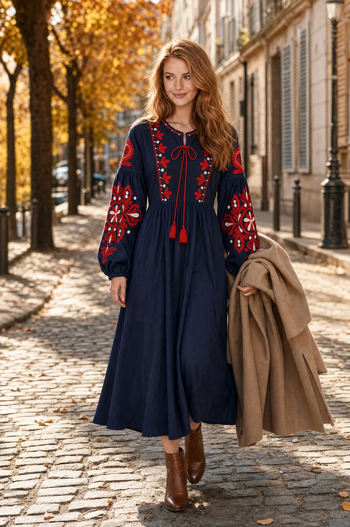Robe bohème brodée bleu marine Constance portée par Julie dans une rue pavée européenne en automne, feuillages orangés, manteau camel sur le bras, ambiance élégante d'automne
