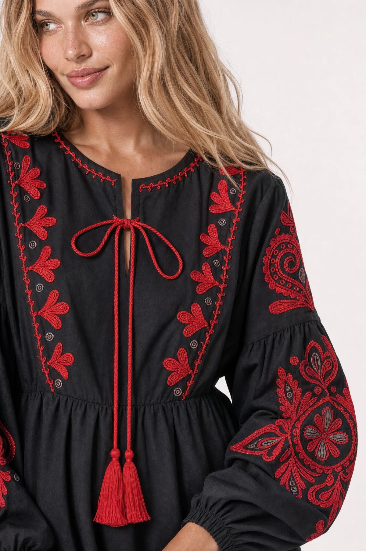Gros plan sur le lien à pompons rouges et la broderie florale rouge façon folklore de la robe bohème brodée noire Agathe, détail du col rond et de la manche longue bouffante brodée portée par Aude