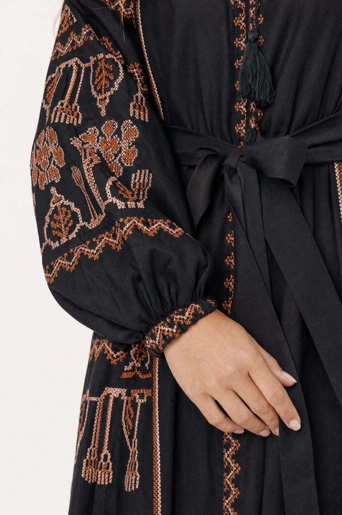 Gros plan sur la broderie ethnique rouille et terracotta et la ceinture nouée à pompons de la robe bohème noire Salomé, détail des manches bouffantes resserrées au poignet et du tissu coton lin