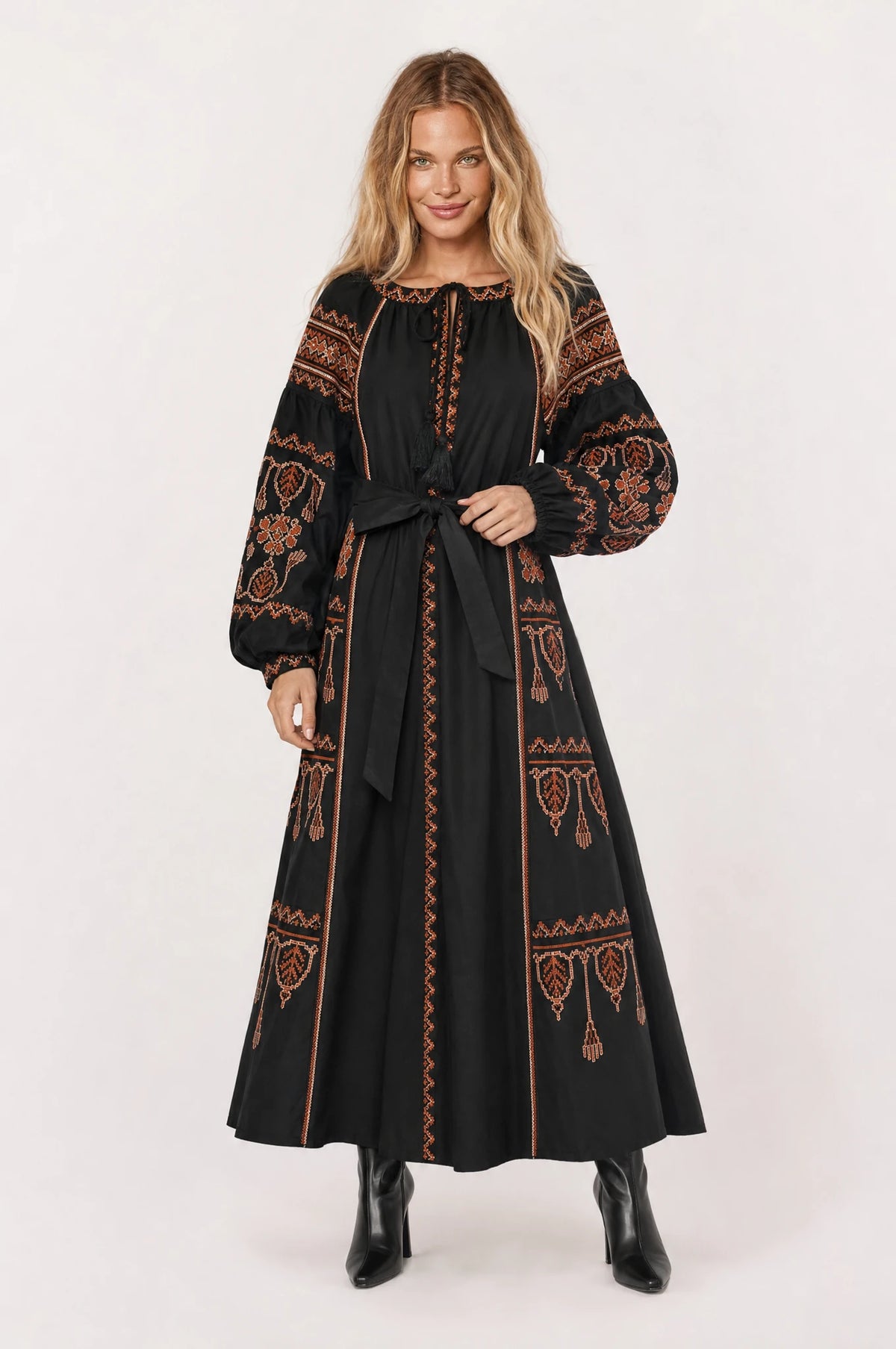 Robe bohème brodée noire Salomé portée de face par Aude cheveux détachés, décolleté rond fermé par un lacet à pompons, broderie ethnique rouille et terracotta sur les manches bouffantes et la jupe, ceinture nouée à la taille, bottines noires, tenue chic automne hiver
