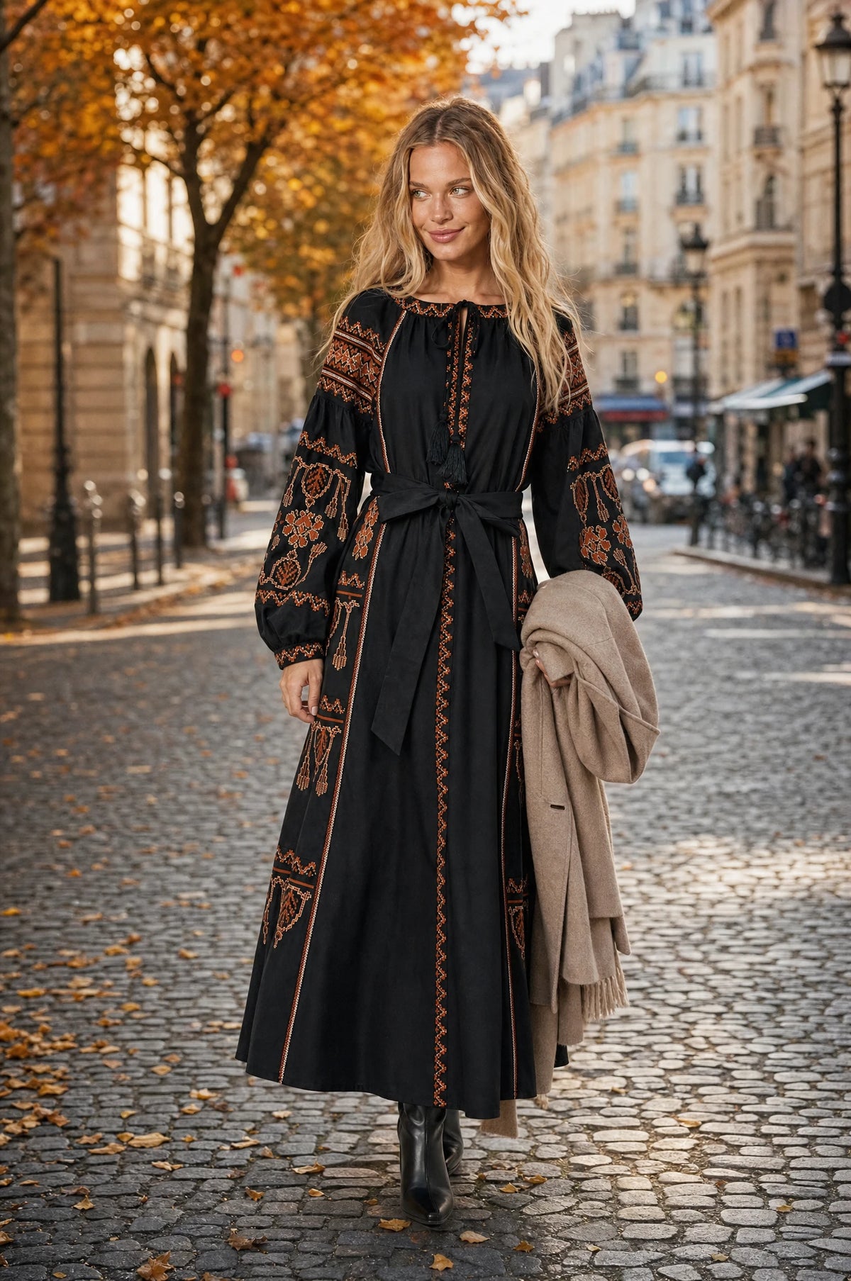 Robe bohème brodée noire Salomé portée par Aude dans une rue parisienne chic en automne, feuillages dorés, manteau camel porté sur le bras, bottines noires, ambiance élégante de journée d'automne