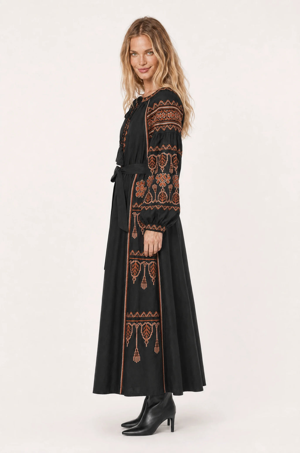 Robe bohème brodée noire Salomé vue de profil portée par Aude cheveux détachés, silhouette évasée coupe empire, broderie ethnique rouille et terracotta, jupe longue jusqu'aux chevilles, ceinture nouée à la taille, bottines noires à talon