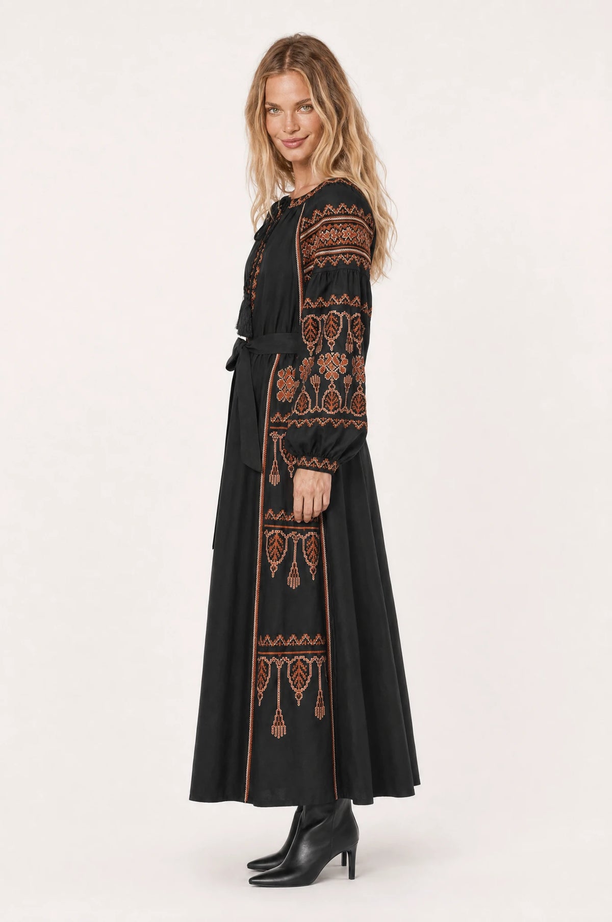 Robe bohème brodée noire Salomé vue de profil portée par Aude cheveux détachés, silhouette évasée coupe empire, broderie ethnique rouille et terracotta, jupe longue jusqu'aux chevilles, ceinture nouée à la taille, bottines noires à talon