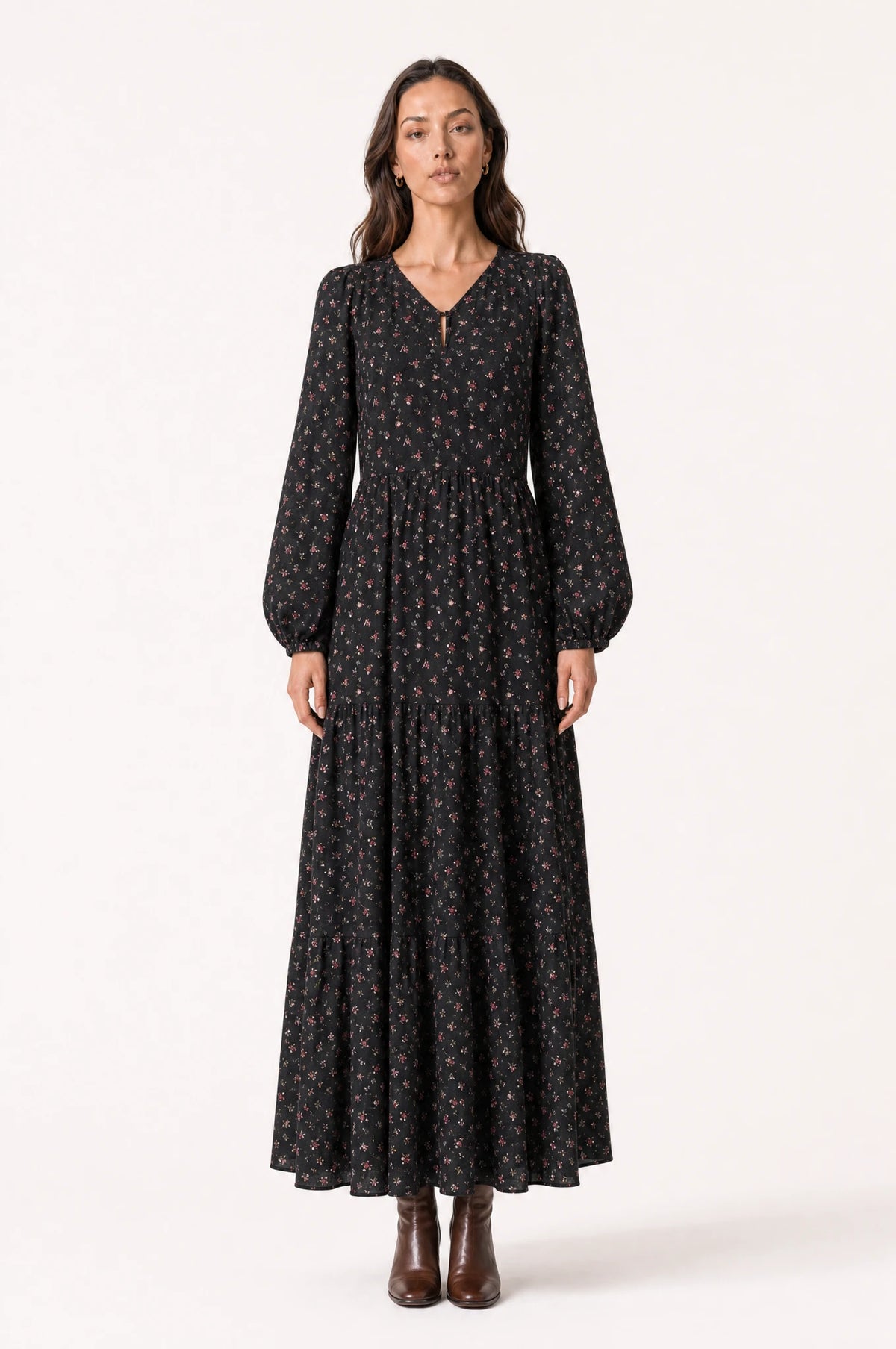 Robe bohème manche longue fleurie noire Pauline portée de face par Nath cheveux détachés, col en V pointu avec bouton keyhole, manches longues bouffantes, taille marquée, jupe évasée à volants, bottines en cuir marron