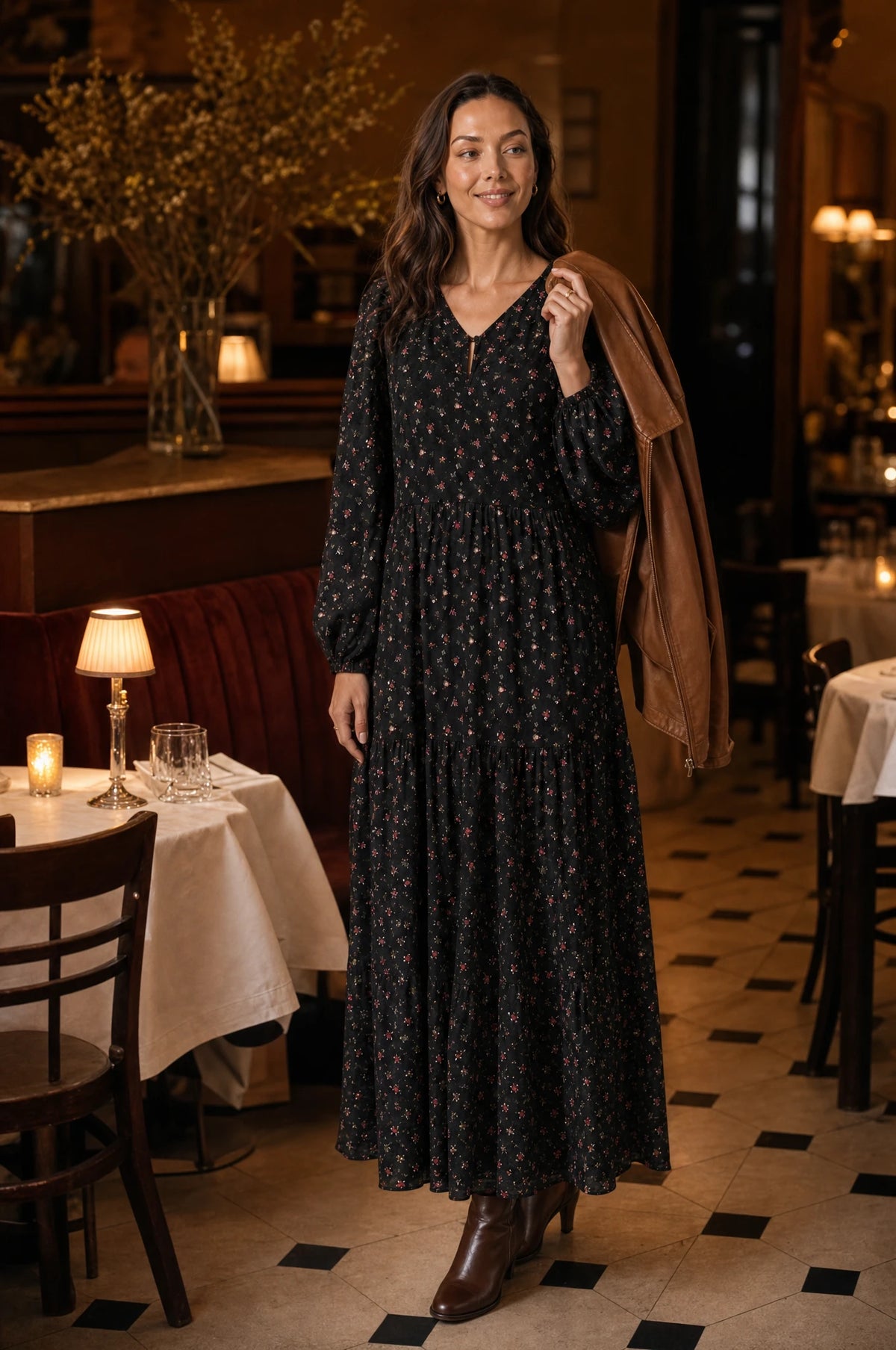 Robe bohème manche longue fleurie noire Pauline portée par Nath dans un restaurant chic en soirée, veste en cuir camel sur l'épaule, lumière chaude des bougies, ambiance conviviale de dîner Image