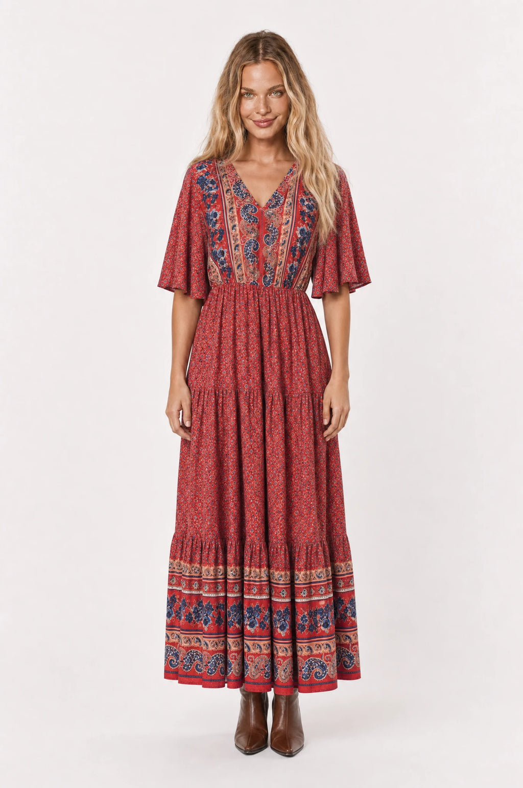 Robe bohème longue fleurie rouge Marguerite portée de face par Aude cheveux détachés, décolleté en V bordure paisley, manches courtes évasées, taille élastiquée, jupe évasée à volants fermée, bottines marron