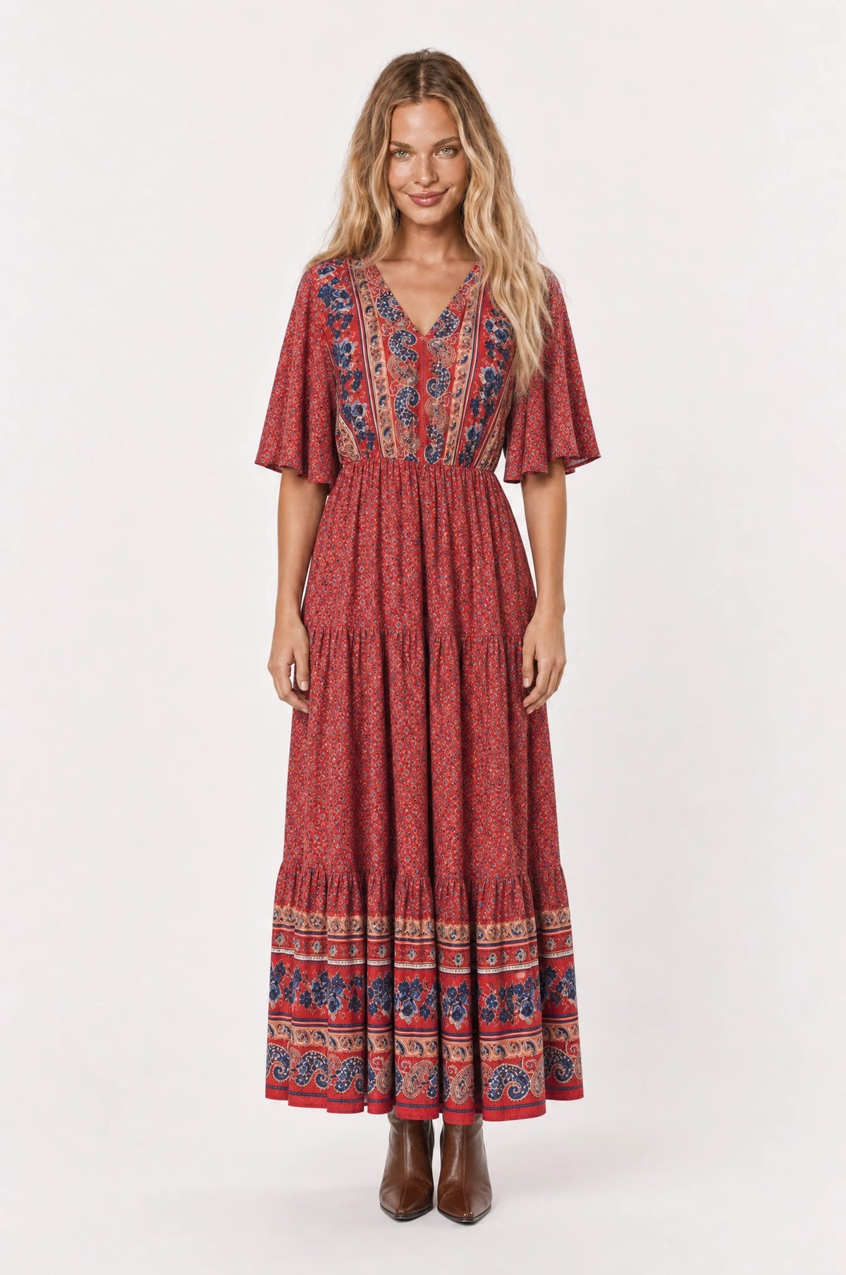 Robe bohème longue fleurie rouge Marguerite portée de face par Aude cheveux détachés, décolleté en V bordure paisley, manches courtes évasées, taille élastiquée, jupe évasée à volants fermée, bottines marron