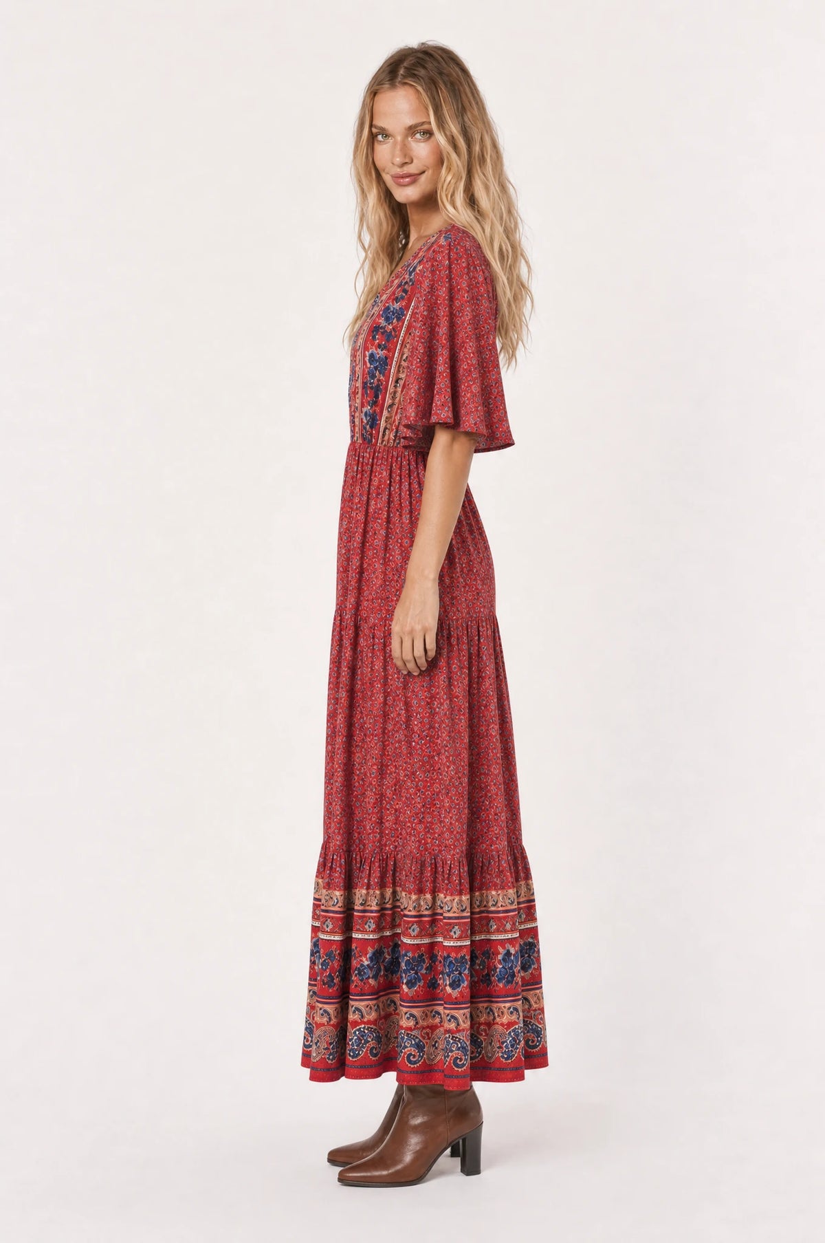 Robe bohème longue fleurie rouge Marguerite vue de profil portée par Aude cheveux détachés, silhouette évasée taille élastiquée, jupe longue jusqu'à la cheville, bottines en cuir marron