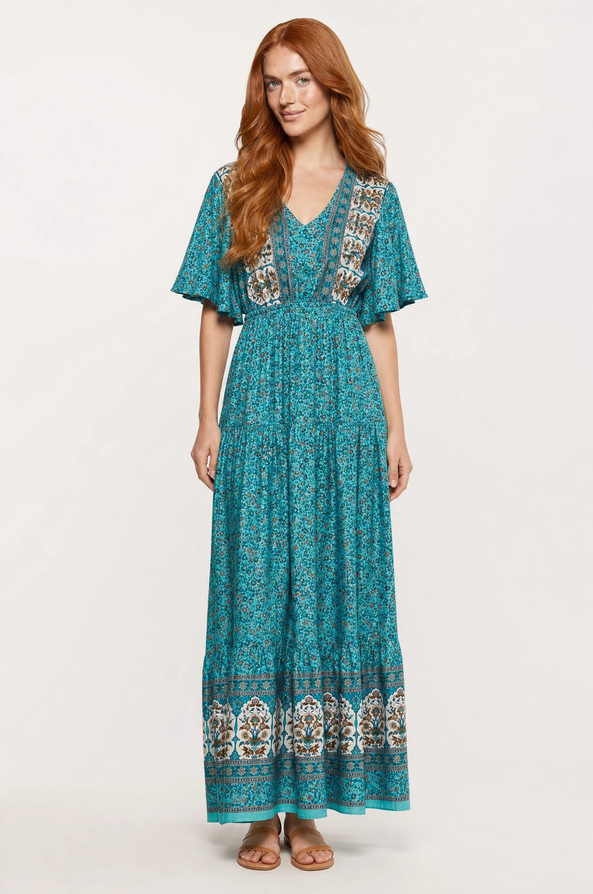 Robe bohème longue fleurie turquoise Céleste portée de face par Julie cheveux roux ondulés détachés, décolleté en V bordure paisley, manches courtes évasées, taille élastiquée, jupe évasée à volants fermée, sandales en cuir