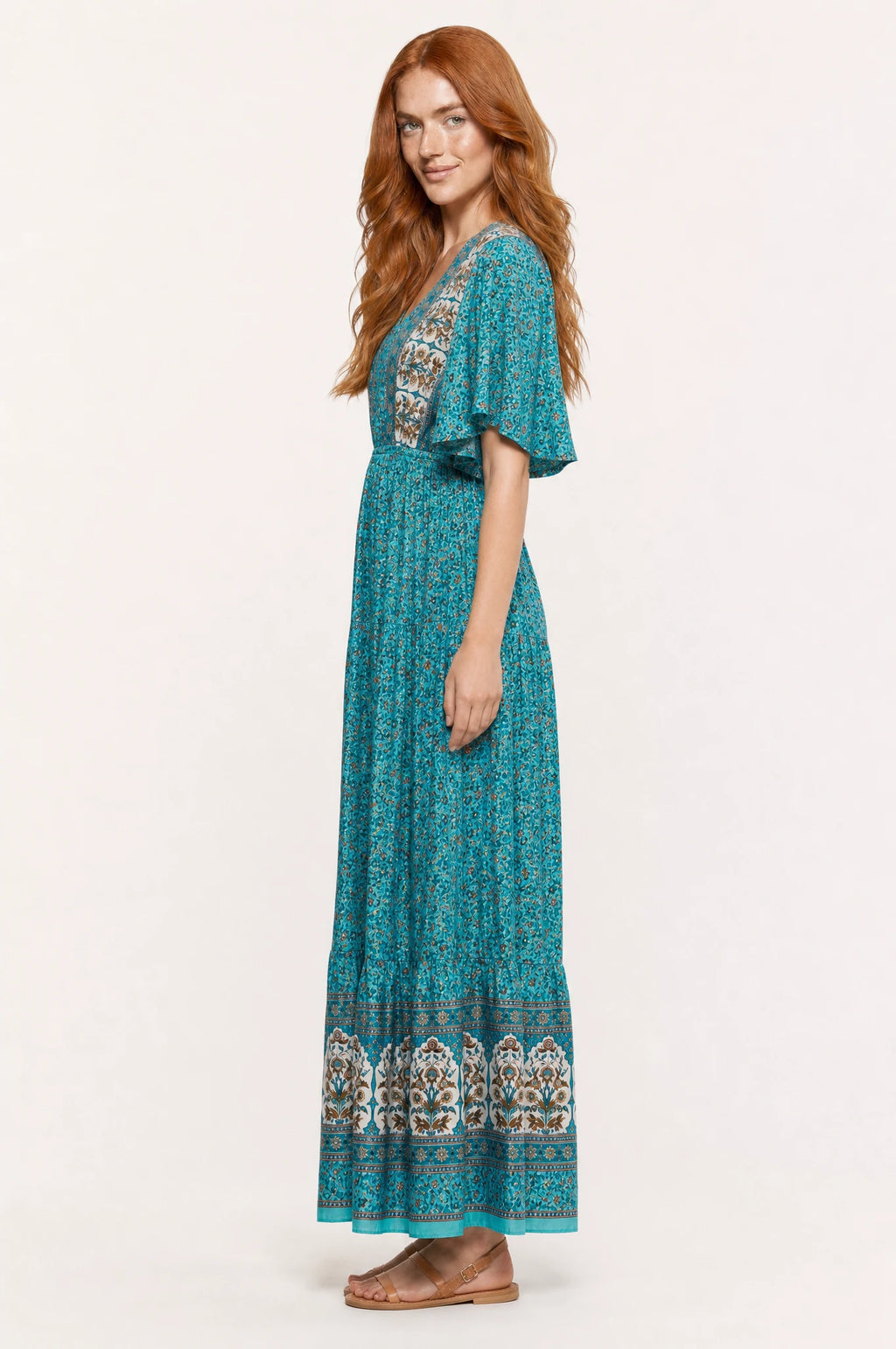 Robe bohème longue fleurie turquoise Céleste vue de profil portée par Julie cheveux roux ondulés détachés, silhouette évasée taille élastiquée, jupe longue jusqu'à la cheville, sandales en cuir marron