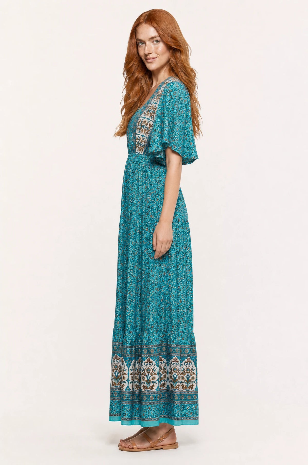 Robe bohème longue fleurie turquoise Céleste vue de profil portée par Julie cheveux roux ondulés détachés, silhouette évasée taille élastiquée, jupe longue jusqu'à la cheville, sandales en cuir marron