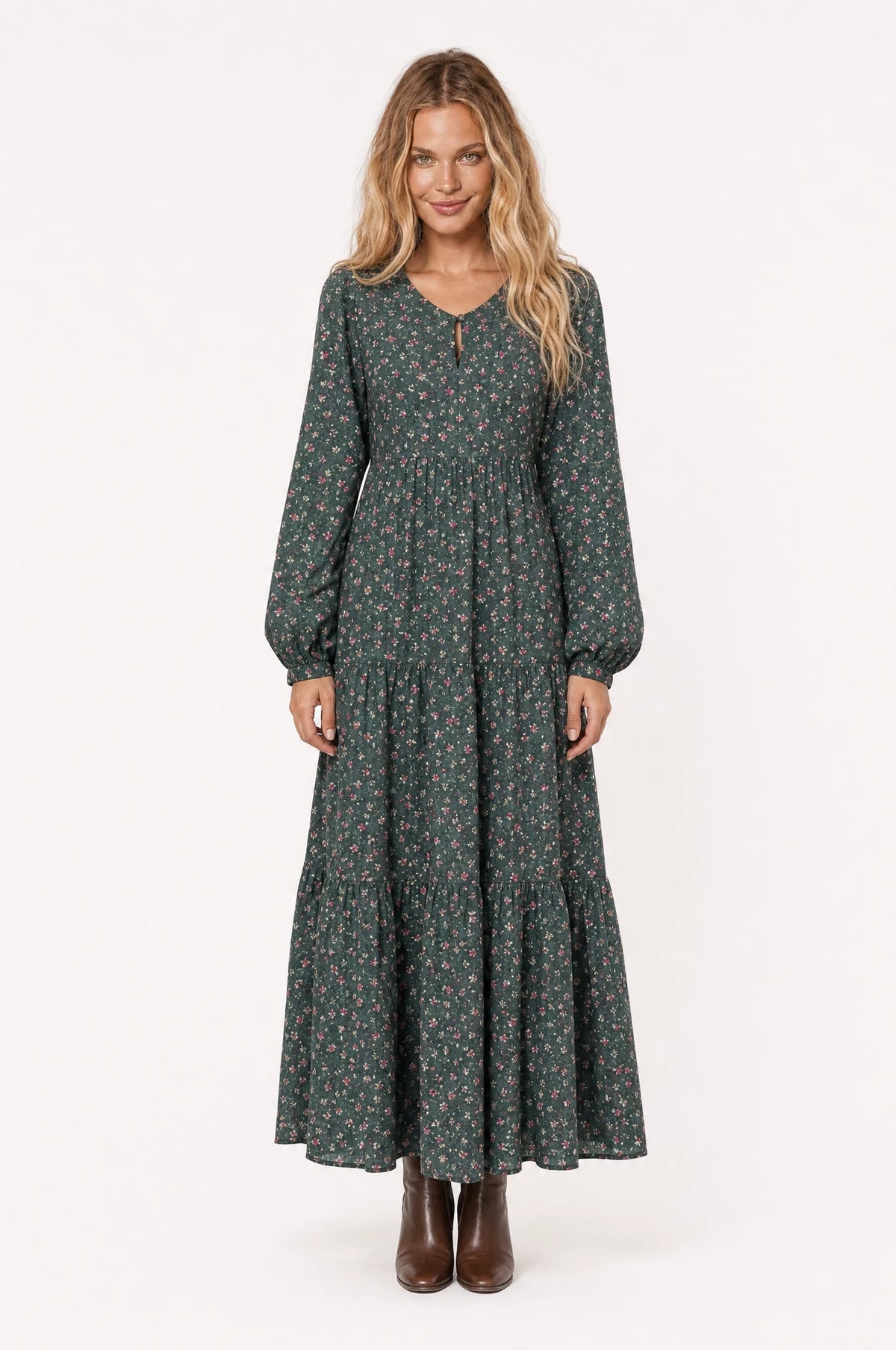 Robe bohème champêtre fleurie verte Roxane portée de face par Aude cheveux détachés, col en V avec bouton keyhole, manches longues bouffantes, taille marquée, jupe évasée à volants, bottines en cuir marron