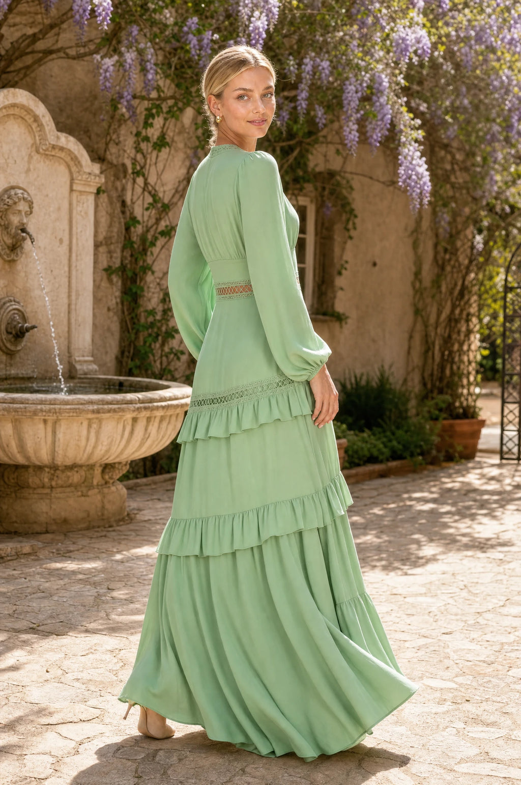 Robe bohème longue vert clair Ondine portée par Sophie dans une cour ensoleillée avec fontaine en pierre et glycine en fleurs, mains libres, regard par-dessus l'épaule, ambiance romantique