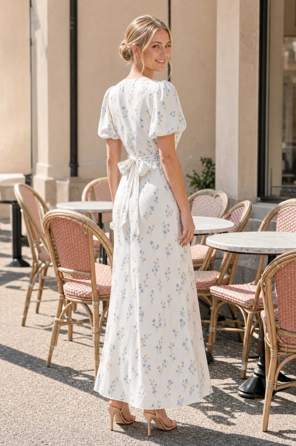 Robe cocktail bleue fleurie Bérénice portée par Aude sur une terrasse de café aux chaises pastel, nœud noué à la taille dans le dos, regard par-dessus l'épaule, mains libres, ambiance décontractée ensoleillée