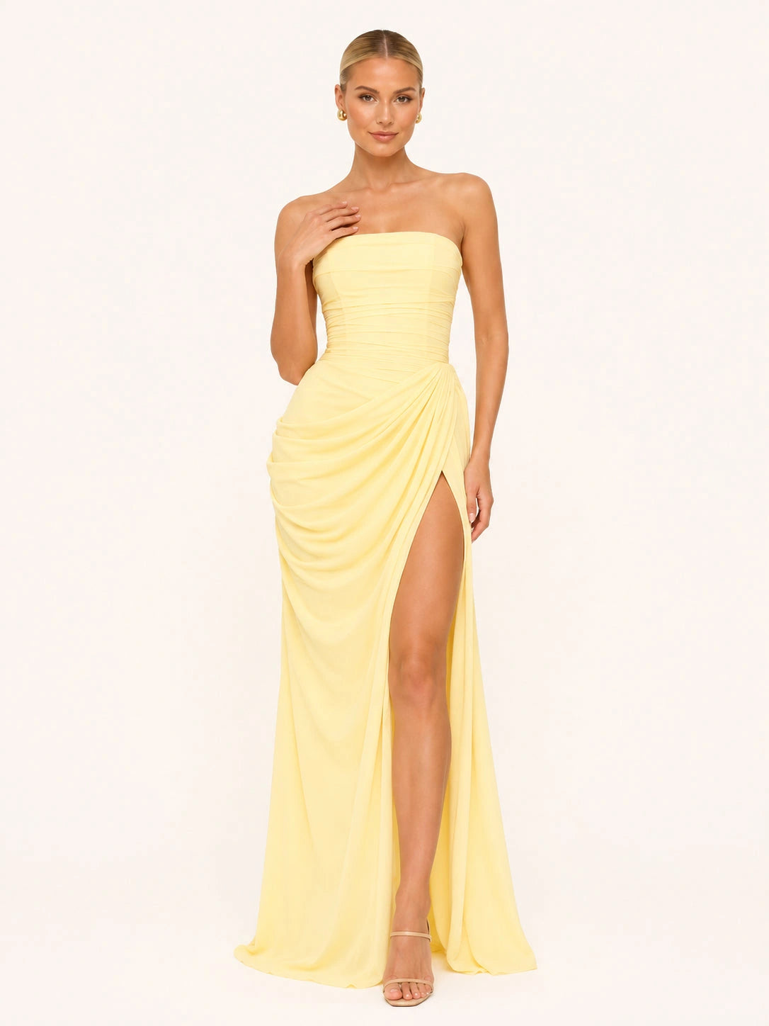 Femme blonde portant une belle robe longue avec un corset bustier jaune pastel. Elle pose sur un fond beige clair. elle a des talons fins et dorés. La robe fendue met en valeur sa jambe.