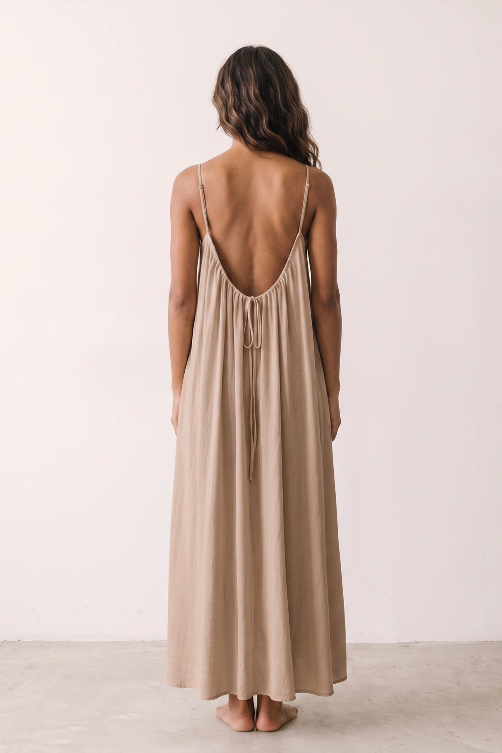 Robe longue casual dos nu Maé couleur Marron clair portée de dos par Camille cheveux détachés, fines bretelles, dos ouvert arrondi avec petit détail bas du dos et coupe fluide sur fond clair