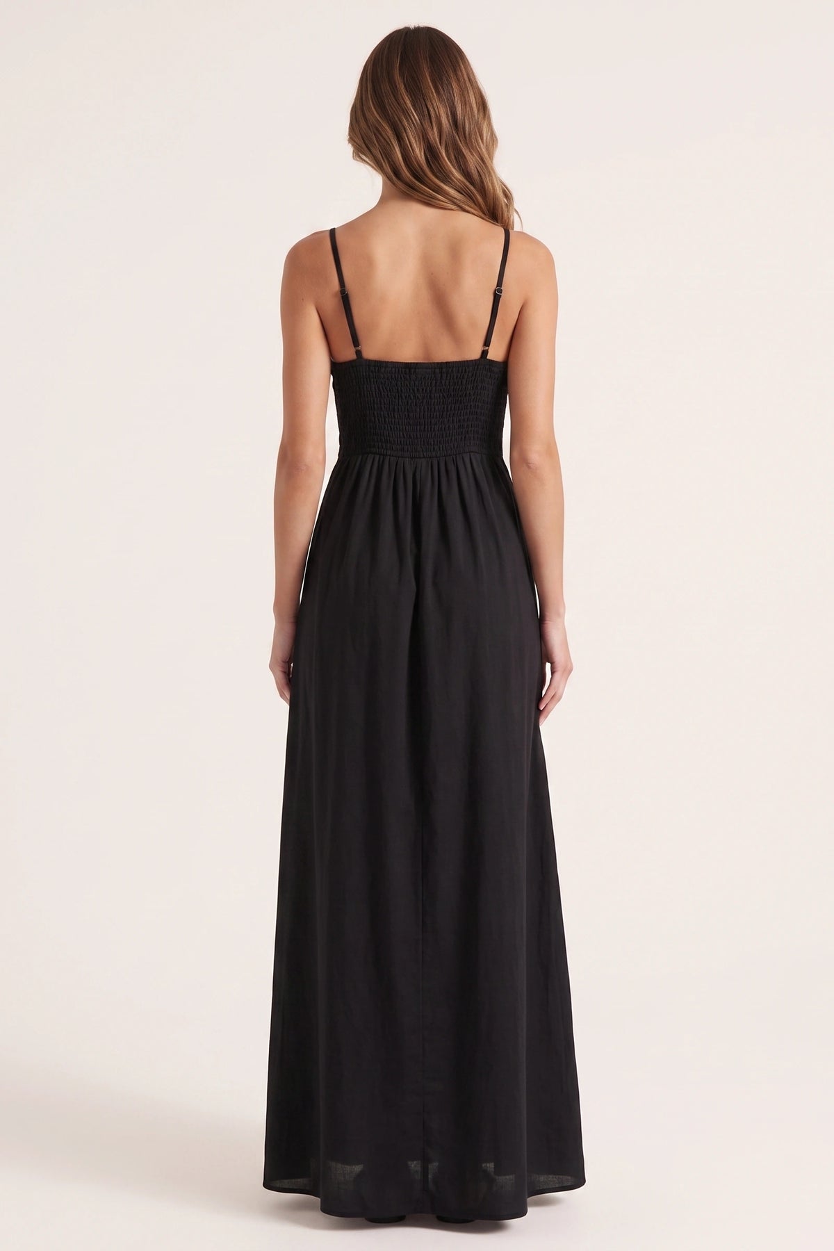 Robe longue casual noire Ava portée de dos par Élena cheveux détachés, dos smocké, fines bretelles et jupe longue fluide sur fond neutre