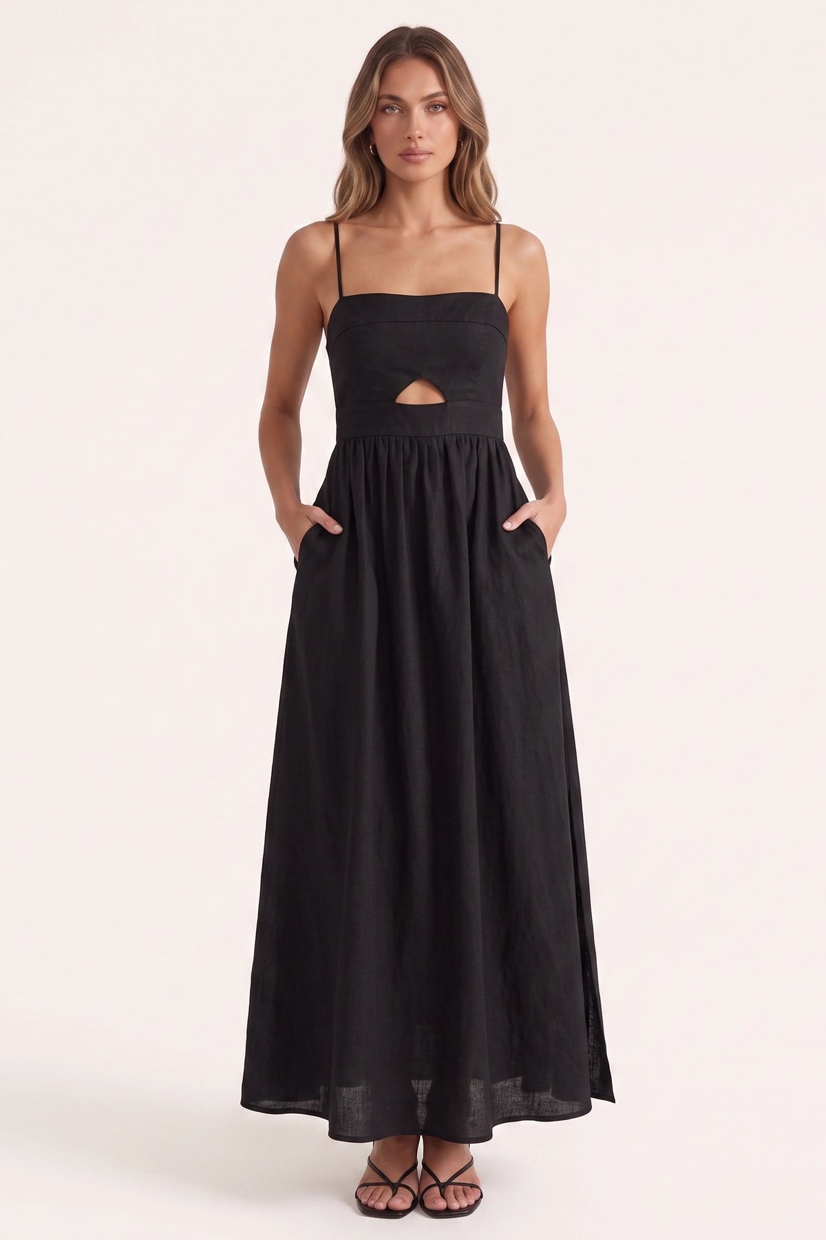 Robe longue casual noire Ava portée de face par le mannequin Élena cheveux détachés, fines bretelles, ouverture triangle, taille froncée, poches et fente côté sur fond clair