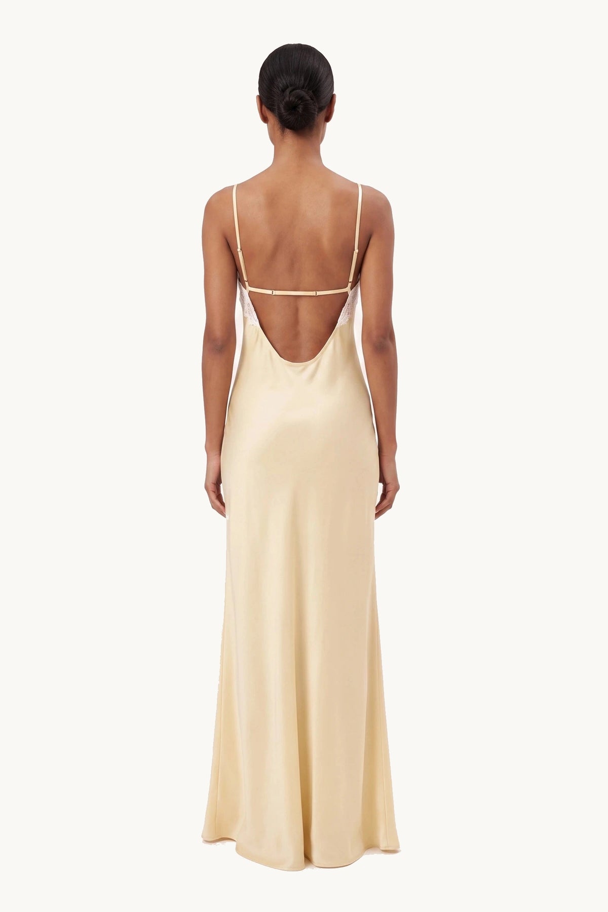 Robe satin dentelle dos nu Alyssa jaune portée de dos par une mannequin métisse sur fond neutre clair, dos nu à fines bretelles et satin fluide