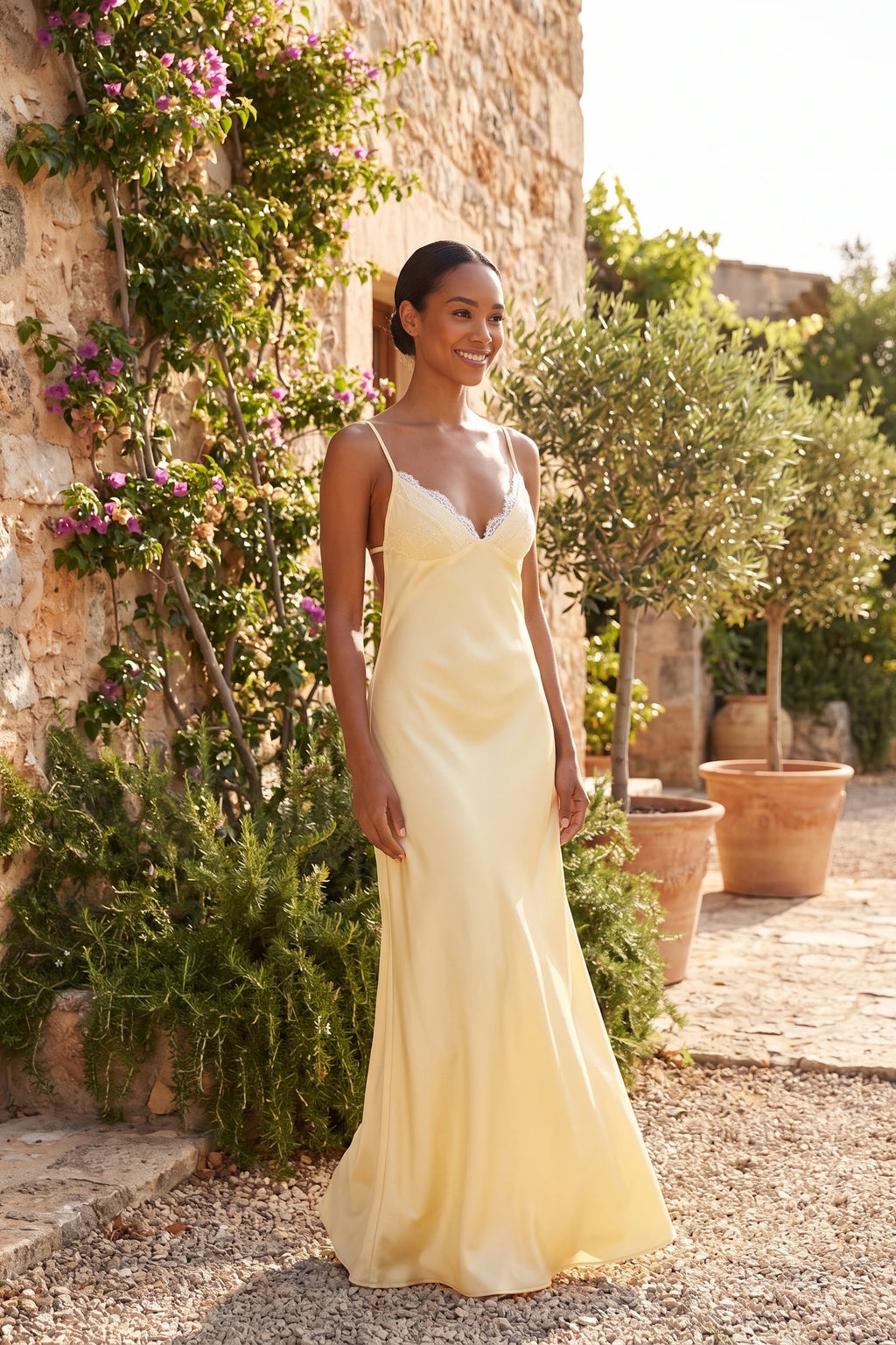 Robe satin dentelle dos nu Alyssa jaune portée par une mannequin métisse dans un jardin méditerranéen, style soirée d’été sans alcool