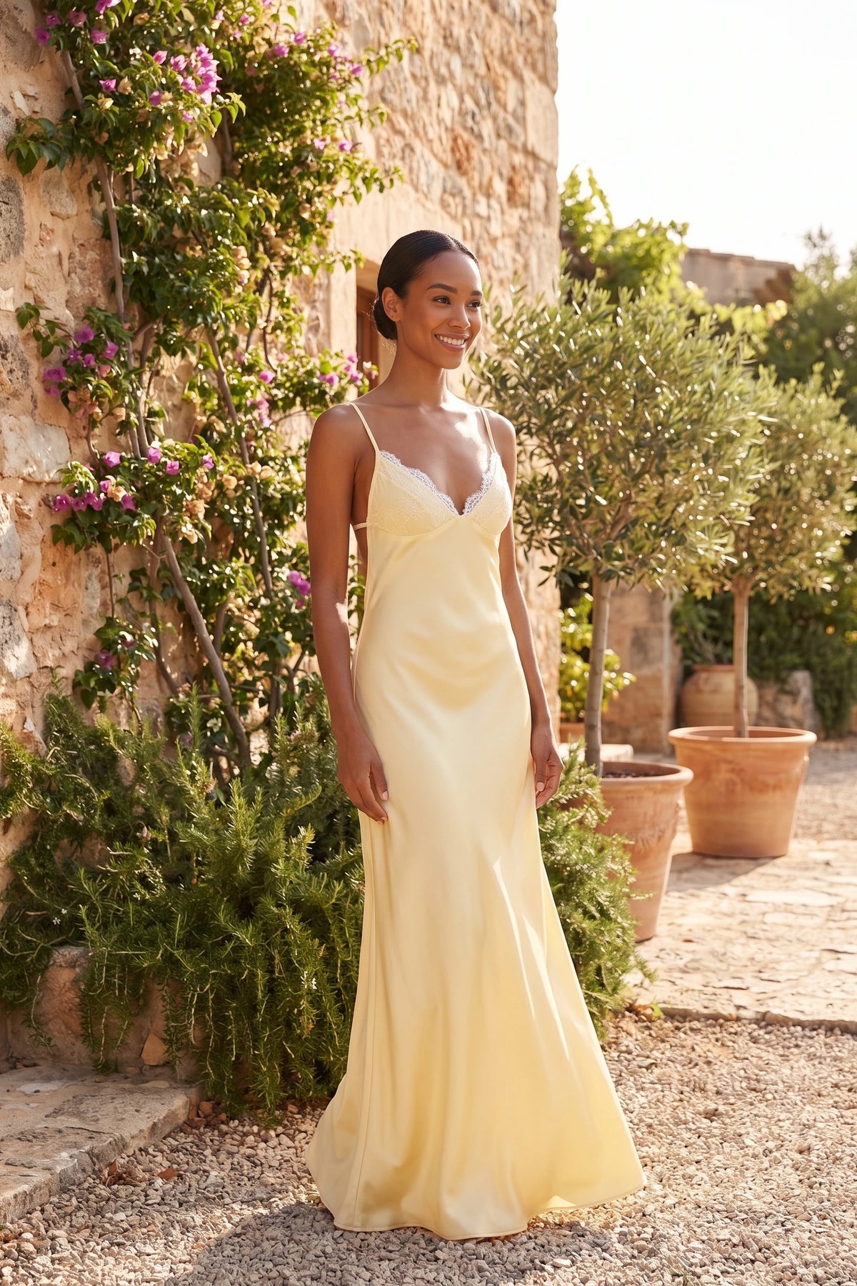Robe satin dentelle dos nu Alyssa jaune portée par une mannequin métisse dans un jardin méditerranéen, style soirée d’été sans alcool