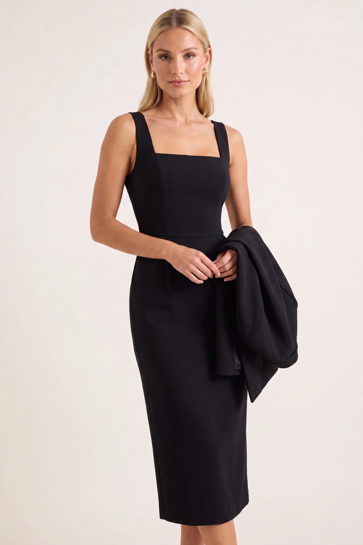 Robe tailleur Alice noire sans cape portée par la mannequin Chloé sur fond beige ivoire fond neutre clair, coupe midi ajustée, bretelles fines et cape noire tenue élégamment sur le bras.