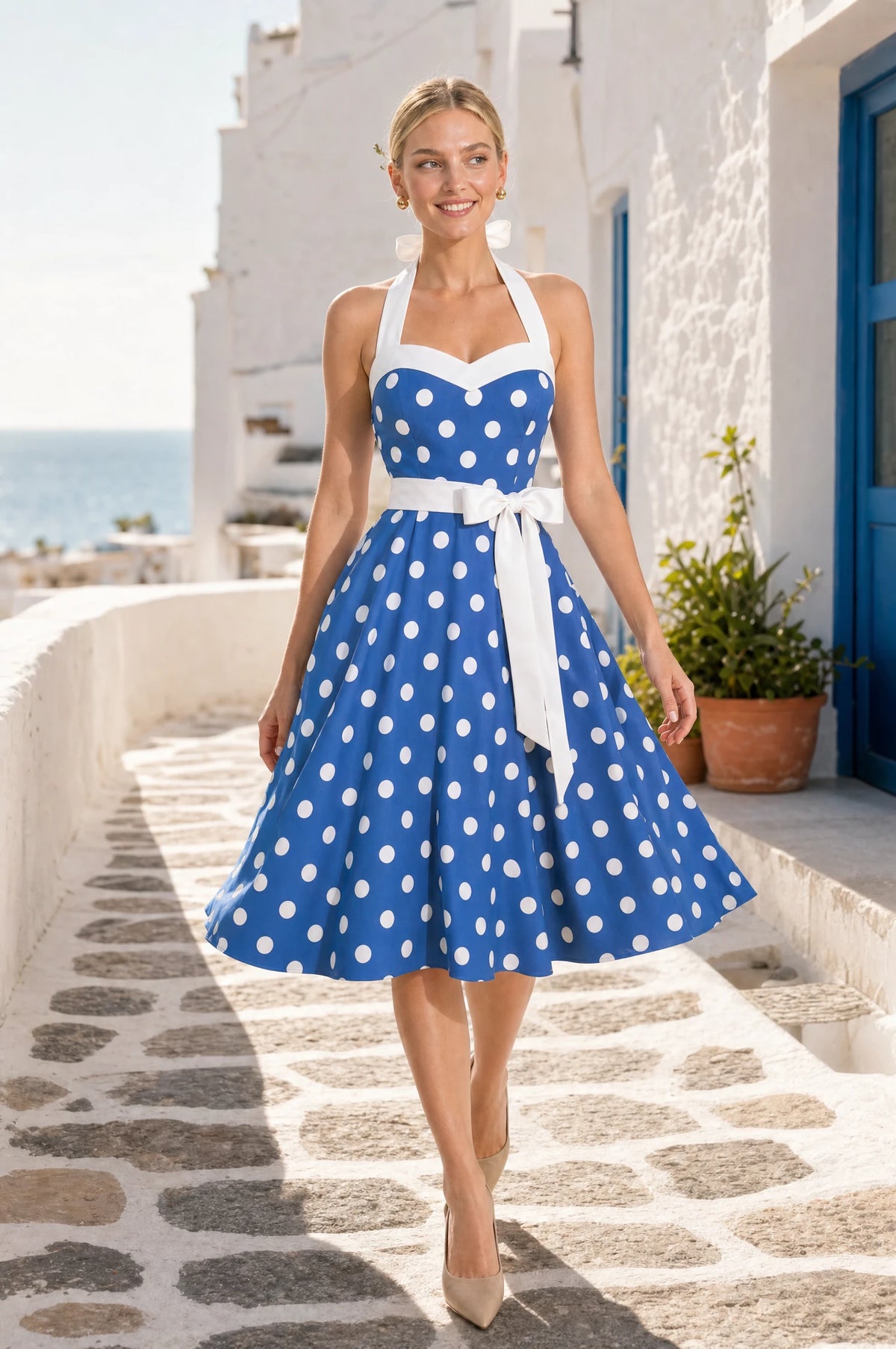 Robe Année 50 bleue à pois Amélie en marche dans une ruelle méditerranéenne aux façades blanches face à la mer, portée par Sophie, regard tourné de côté, ambiance ensoleillée Image