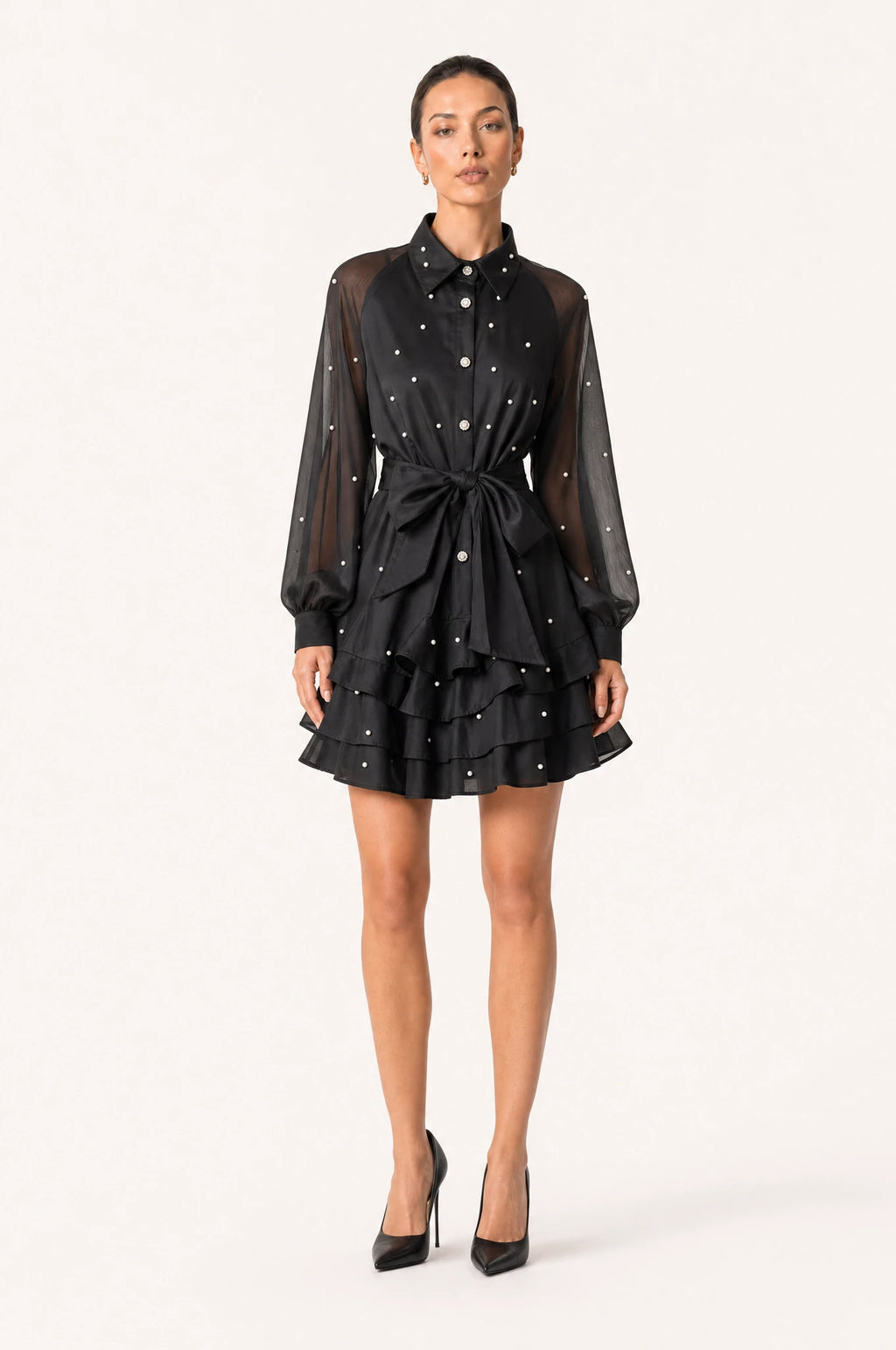 Robe vintage perlée noire Héloïse portée de face par Nath chignon sleek, col chemisier structuré, boutons perlés et strass sur le buste, manches bouffantes en organza semi-transparent, ceinture nouée à la taille, jupe courte à volants superposés