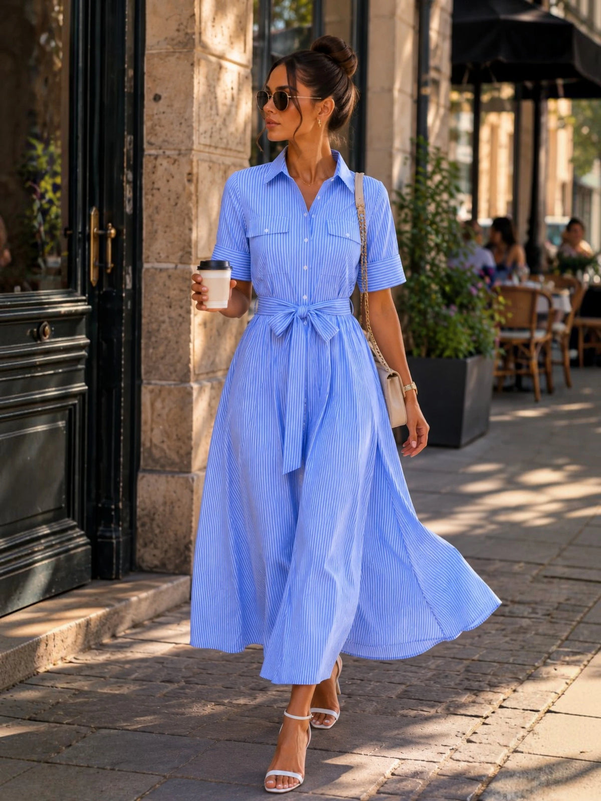 Femme marchant en ville avec une robe chemise longue bleu ciel à rayures fines, jupe fluide en mouvement et coupe élégante, parfaite pour une tenue de ville raffinée et tendance.