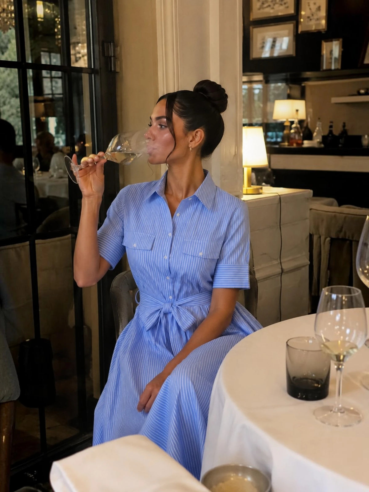 Femme élégante portant une robe chemise longue bleu ciel à fines rayures blanches dans un restaurant raffiné, mettant en valeur une silhouette féminine, une taille ceinturée et un style sophistiqué pour dîner ou événement habillé. Image
