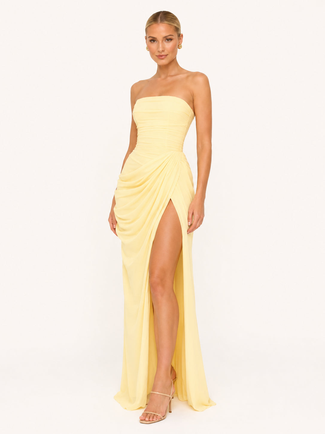 Robe longue bustier jaune pastel, portée par une femme blonde sur un fond beige très clair. La robe fendue laisse apparaître sa jambe.
