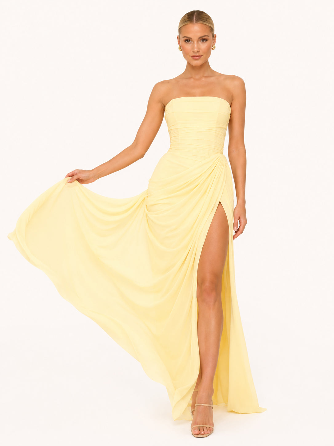 La femme blonde porte une robe bustier longue avec une fente haute de couleur jaune pastel. Elle pose sur un fond beige très clair . Elle tient dans sa main la robe, laissant découvrir sa jambe avec élégance.