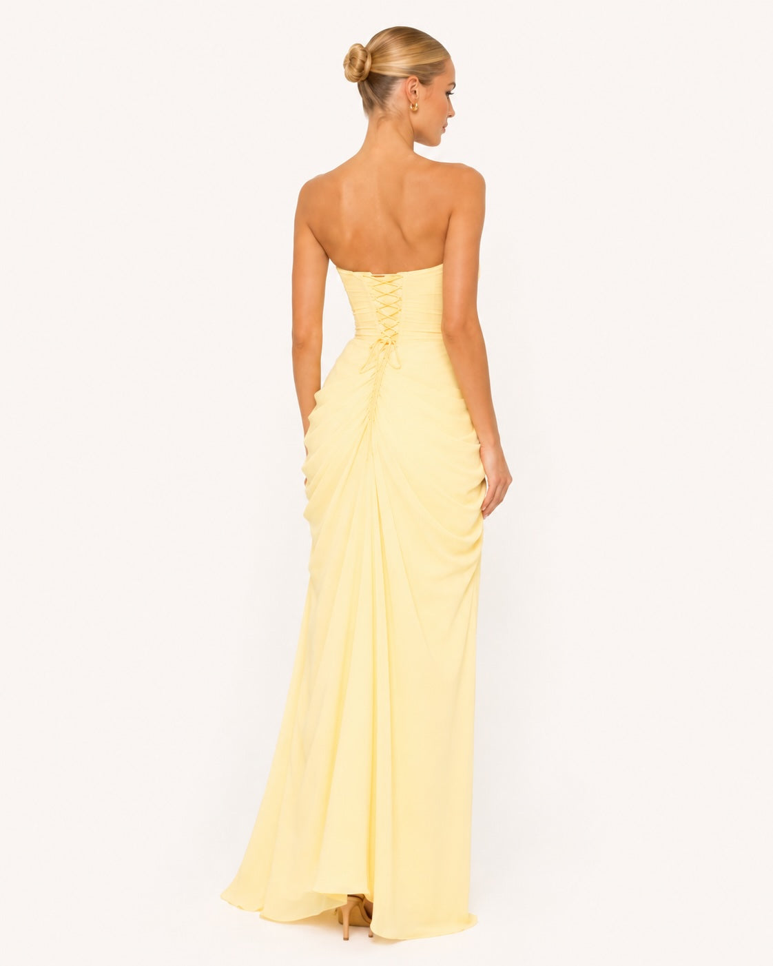 Robe longue élégante jaune pastel portée par une femme posant de dos sur un fond clair. Le corset est lacé dans le dos, le tissus fluide apporte un joli drapé au niveau des hanches.