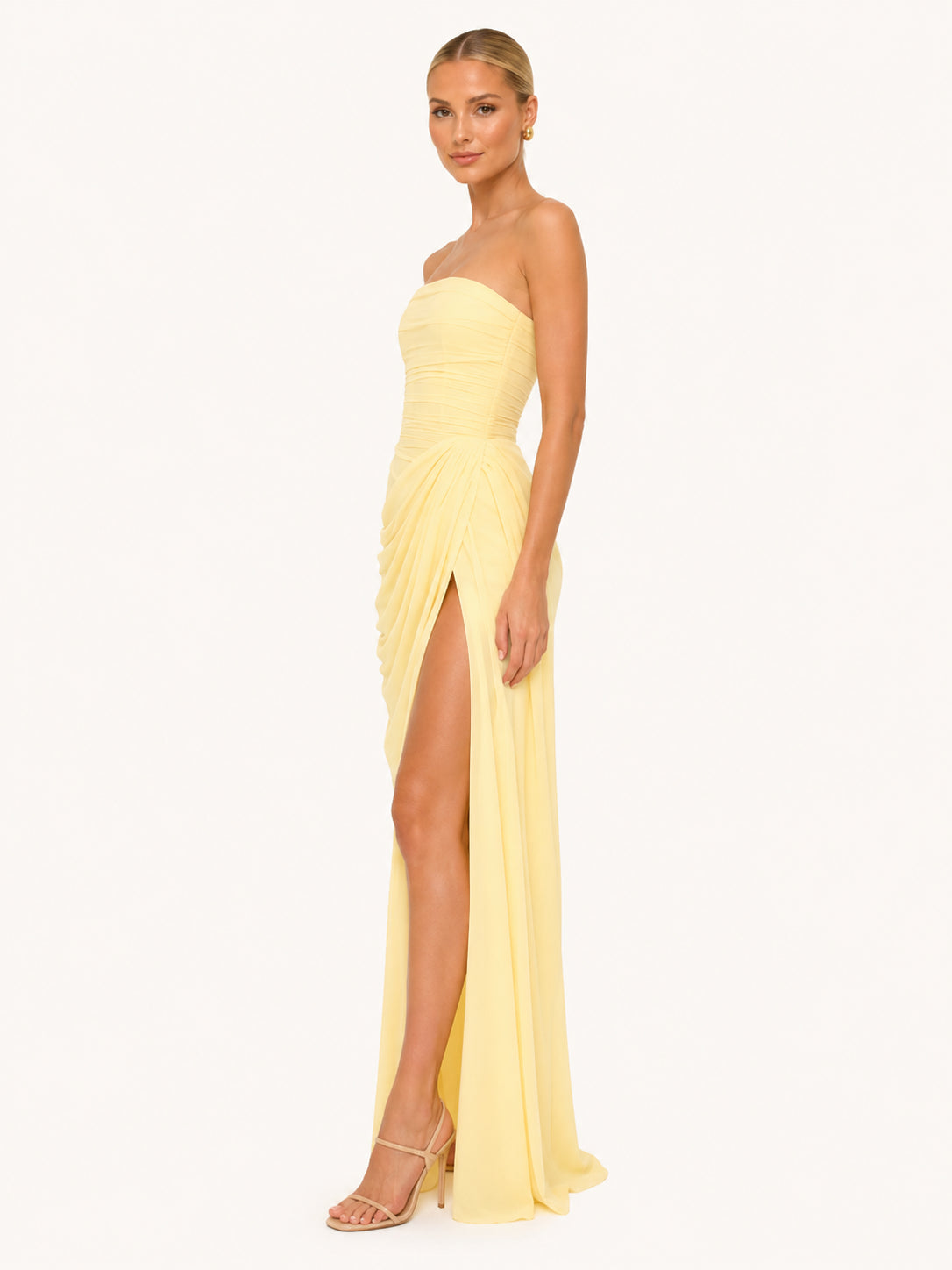 Femme posant de profil sur un fond clair. Elle porte une belle robe longue fendue jaune pastel. Le corset en forme de bustier sculpte sa taille, et le tissu fluide apporte de la légèreté à la robe drapée.