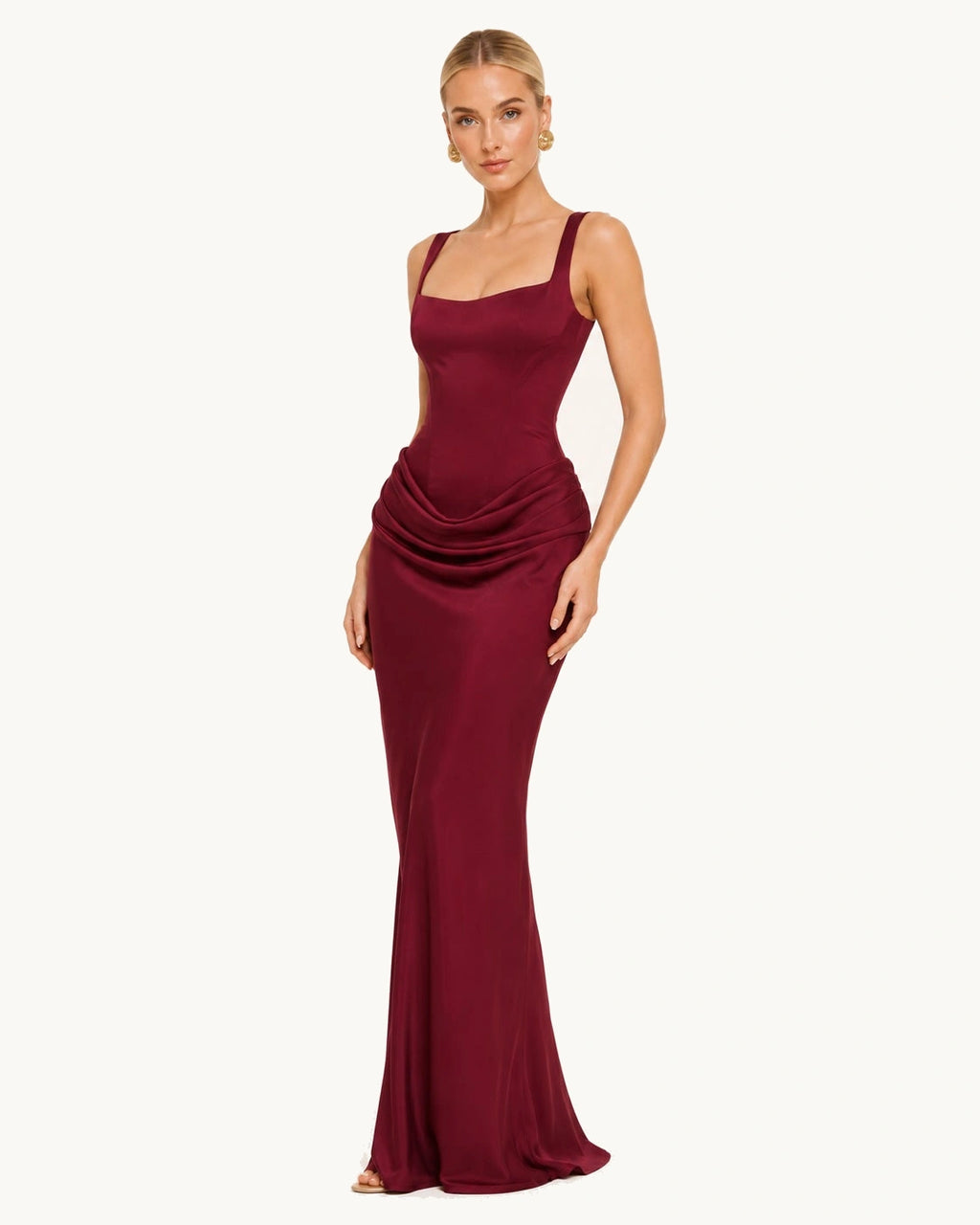 La femme blonde porte une robe longue décolleté bordeaux. La robe est satinée, drapée en niveau des hanches et cintrée à la taille.