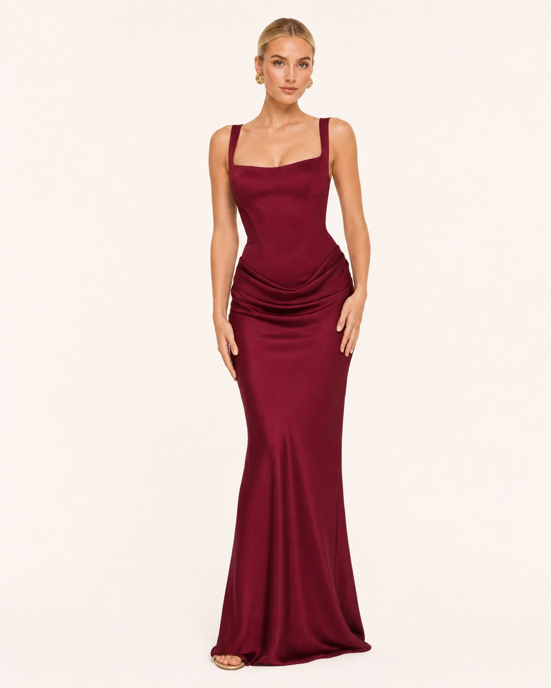 Robe longue bordeaux élégante portée par une femme blonde, vue de face. Coupe cintrée à la taille avec un joli effet drapé, puis évasée sur la longueur. Idéal pour une soirée, ou un évènement important.